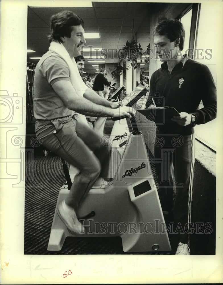 1983 Press Photo Mike Duwa exercises on a Lifecycle with coach Bill Evashevski- Historic Images