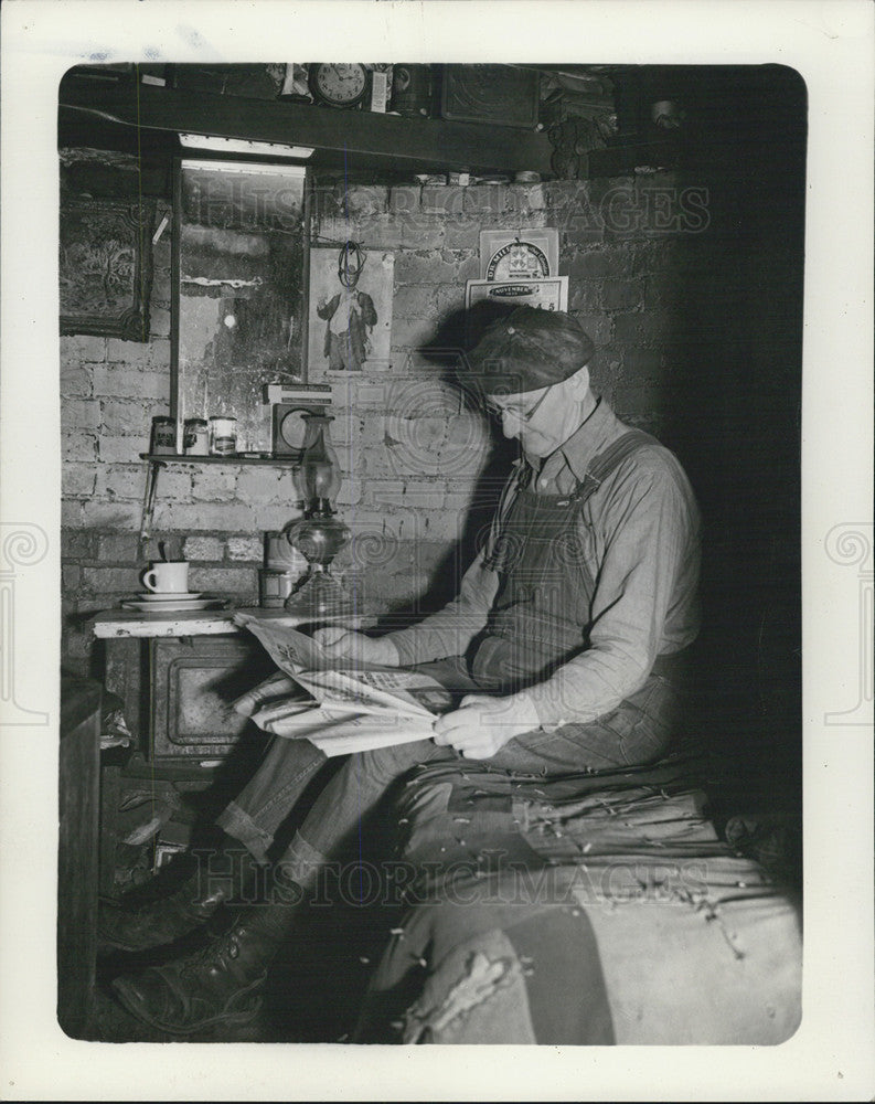 Press Photo Man Reading Newspaper In Small Brick Room Wearing Overalls - Historic Images