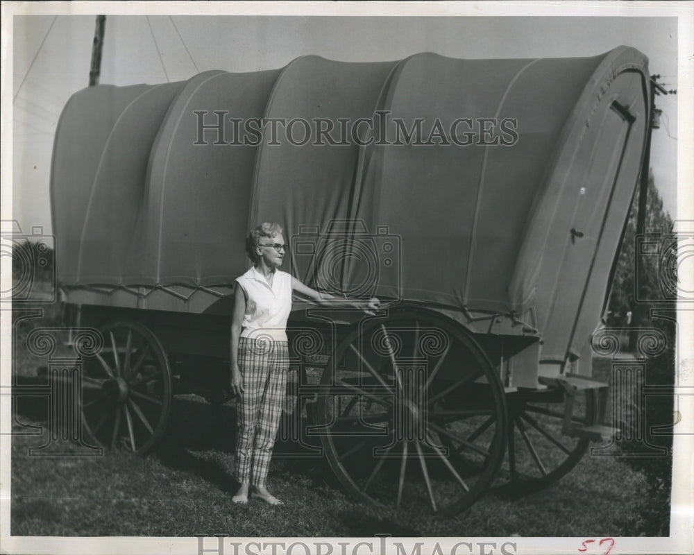 1964 An old lady standing besides a covered wagon - Historic Images