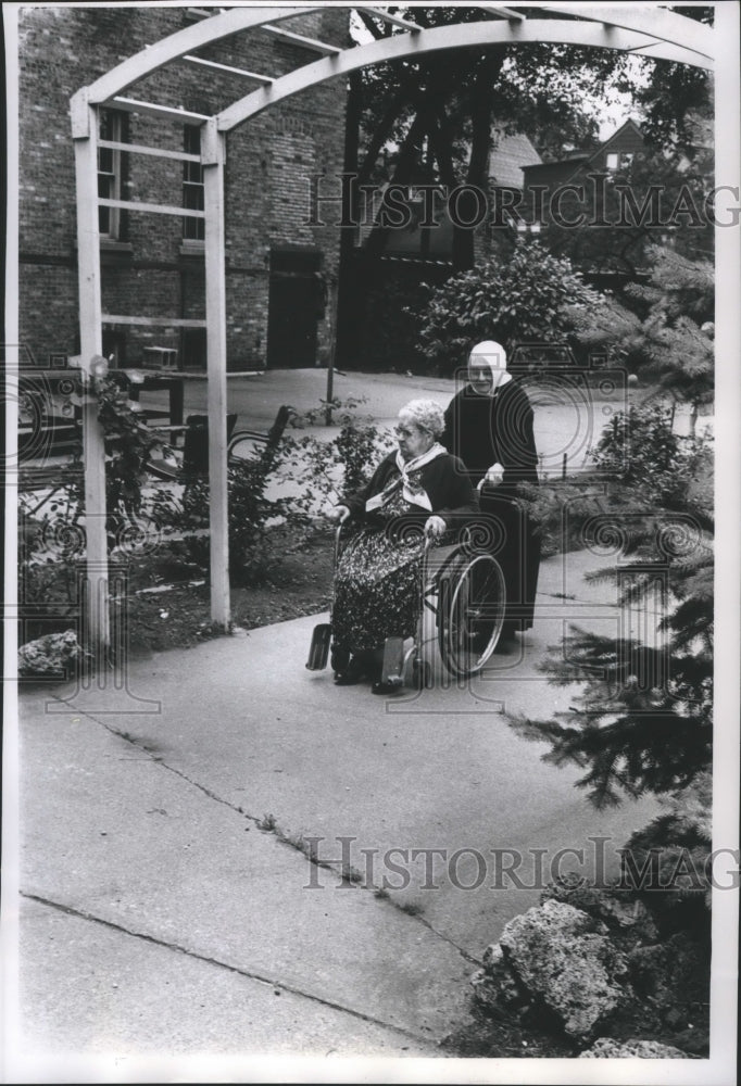 1963 Press Photo Sister Charlotte wheels Mrs. Elizabeth Ferris in a courtyard - Historic Images