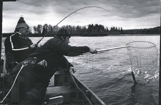 Press Photo One man catches a fish while another nets while fishing from a boat - Historic Images