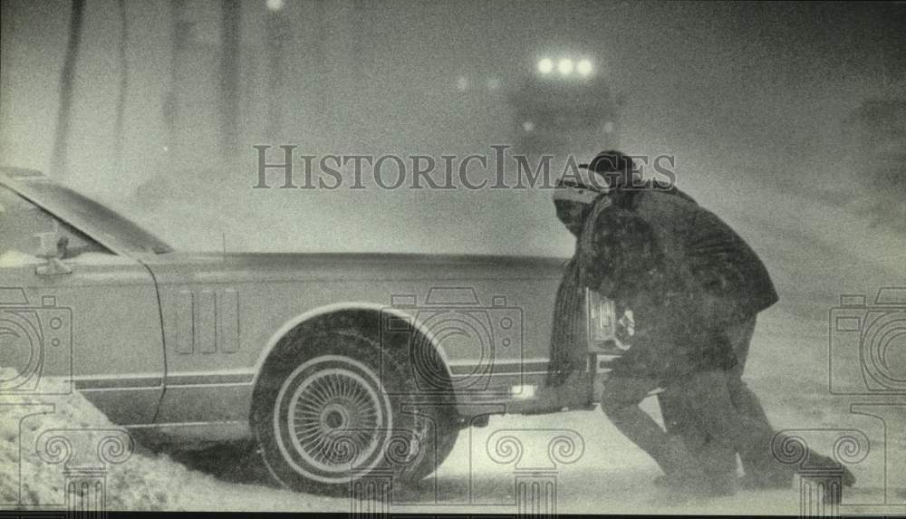 1982 Couple tries to help push car into driveway during snowstorm - Historic Images