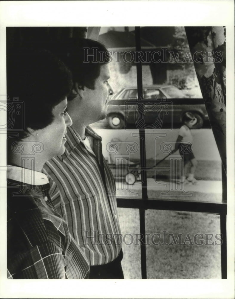 1980 Paul and Catherine Patterson watch child through window. - Historic Images