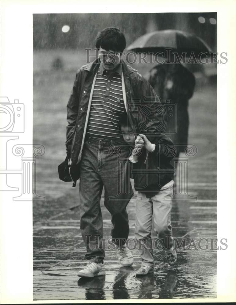 1990 Joe Okamoto peeks from under father&#39;s coat on rainy morning. - Historic Images