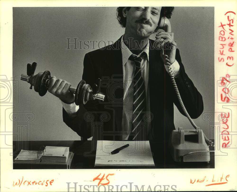 1984 Press Photo Suit-wearing Man lifts Weights while on the Telephone- Historic Images