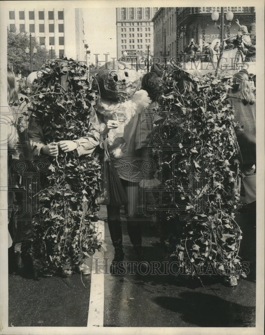 1972 Carnival maskers two bushes walk a pumpkin head down street. - Historic Images