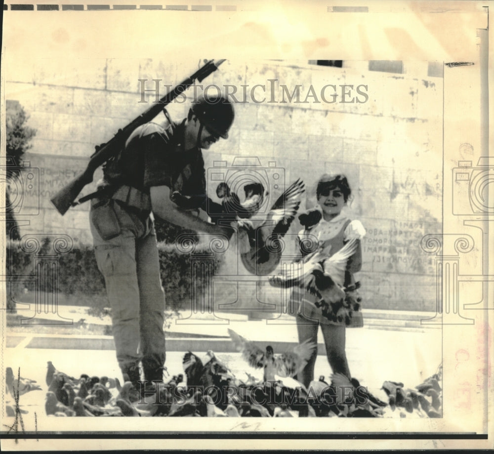 1974 Combat-clad Greek soldier toys with pigeons with child watching - Historic Images