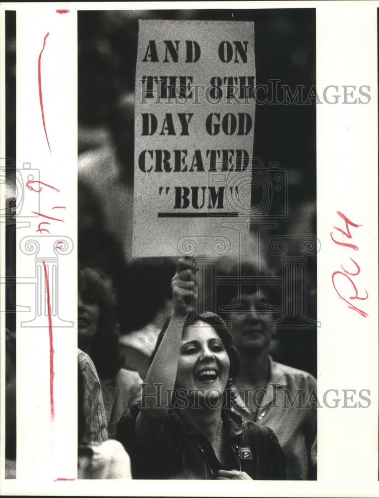 1983 Press Photo Women holds &quot;and on the 8th day God created &#39;BUM&#39;&quot; sign - Historic Images