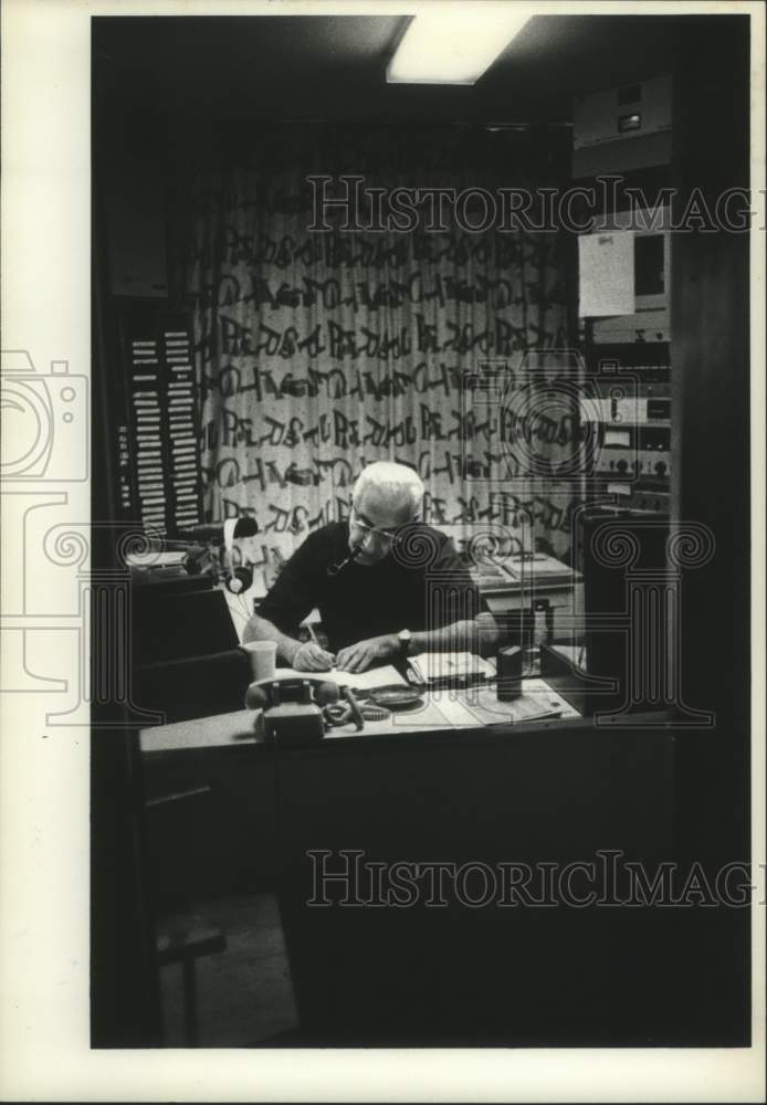 1977 Father Angelers Nitzer sitting at desk with pipe - Historic Images