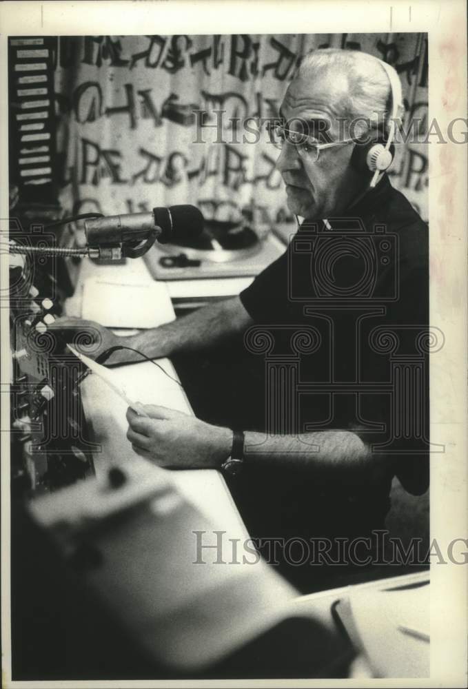 1977  Father Angelers Nitzer sitting at desk with microphone - Historic Images