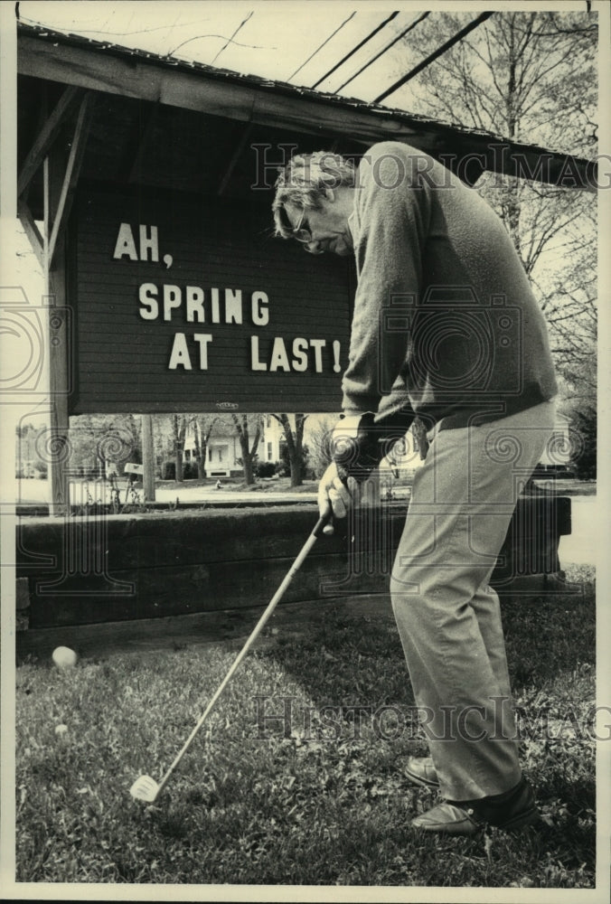 1985 Press Photo Golfer Paul Lassi chips ball in front of 'Spring at Last' sign - Historic Images
